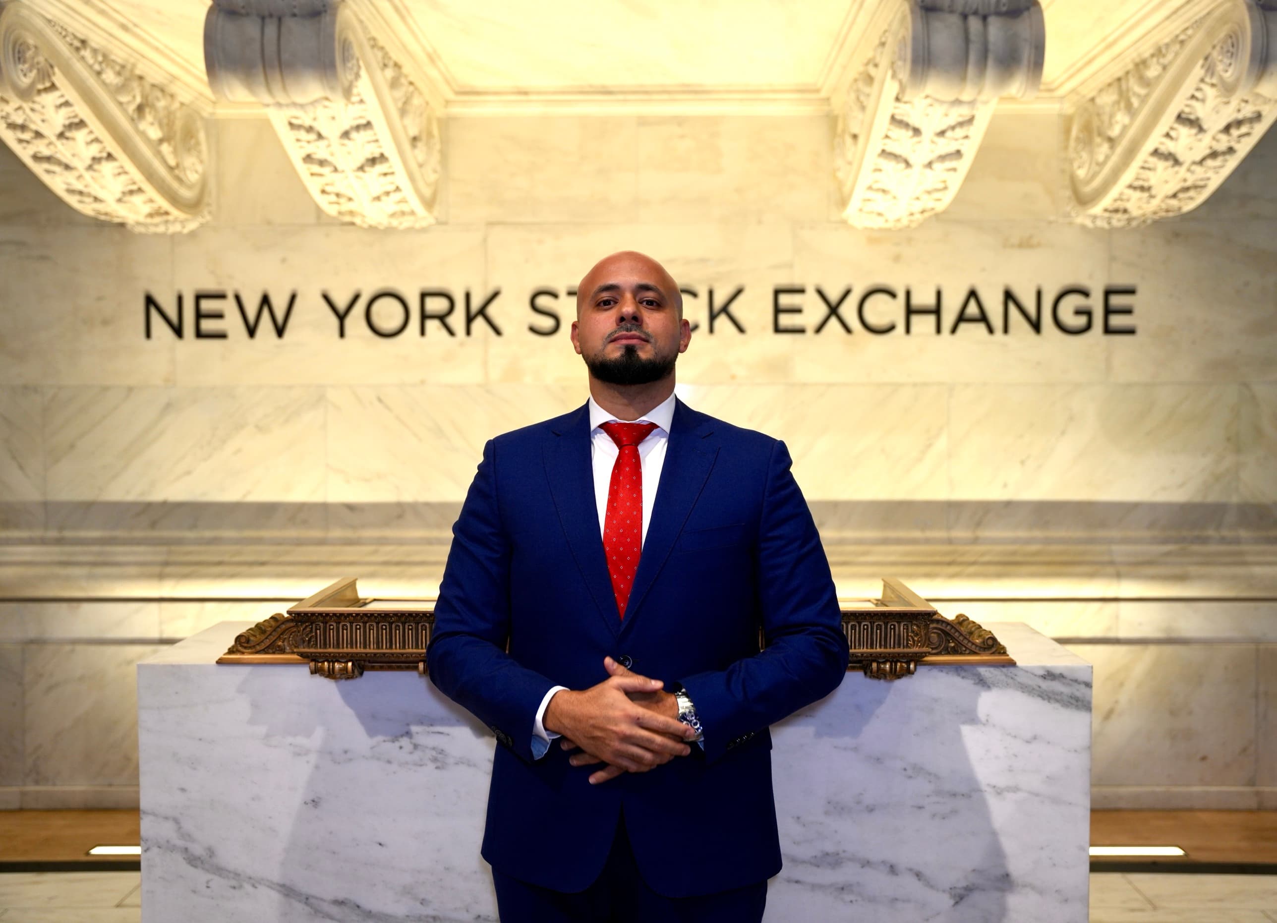 Mohammad Elmoshnib in a navy suit and red tie, standing at the podium in front of the New York Stock Exchange lettering