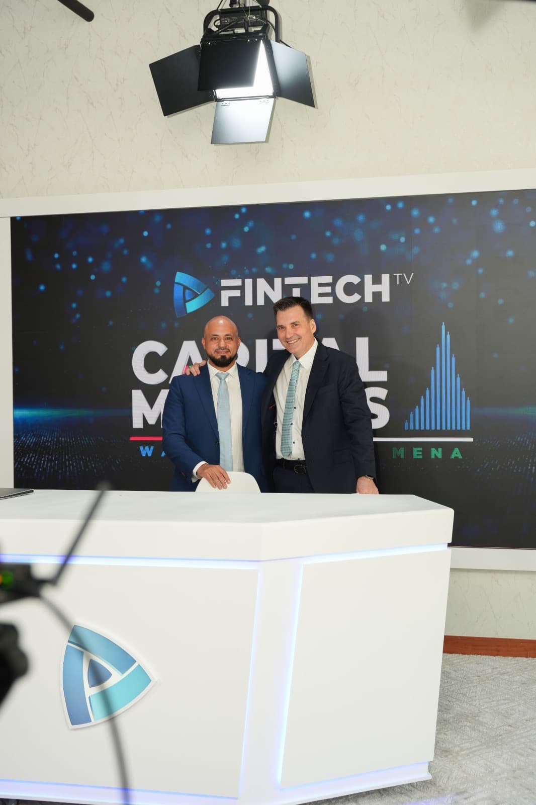 Mohammad Elmoshnib and Troy McGuire stand behind the presenter's desk in the studio, in front of a screen showing the FINTECH.TV name