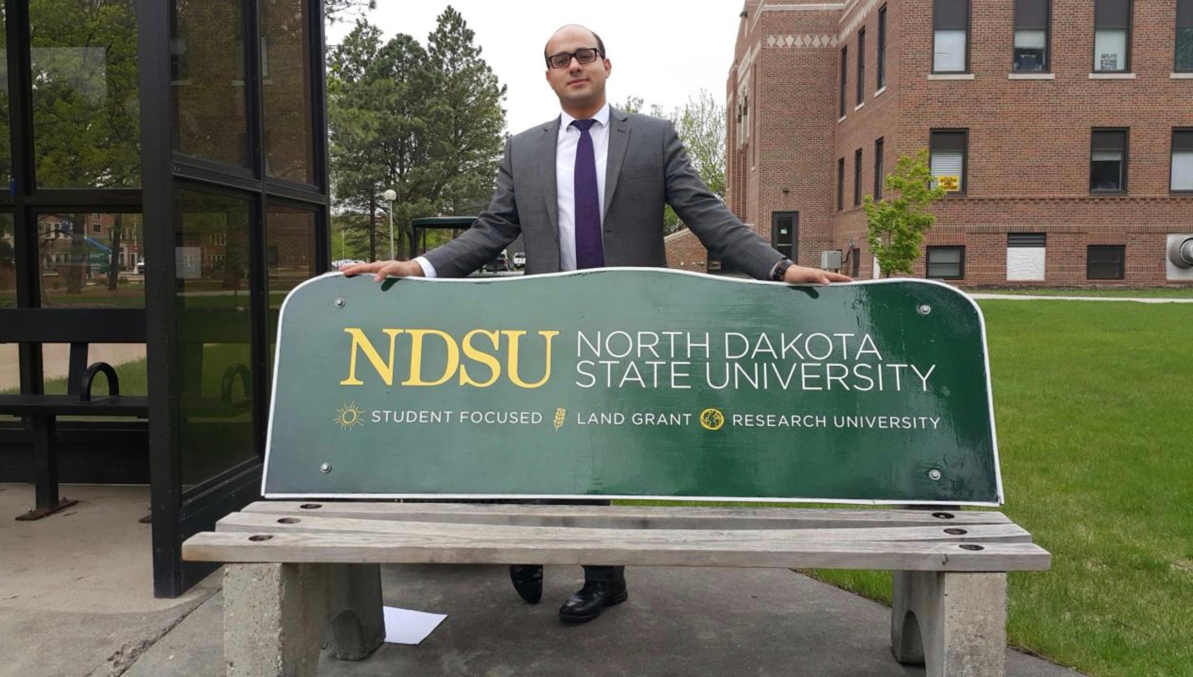Mohammad Elmoshnib standing behind a bench that carries the North Dakota State University name, on the university's campus