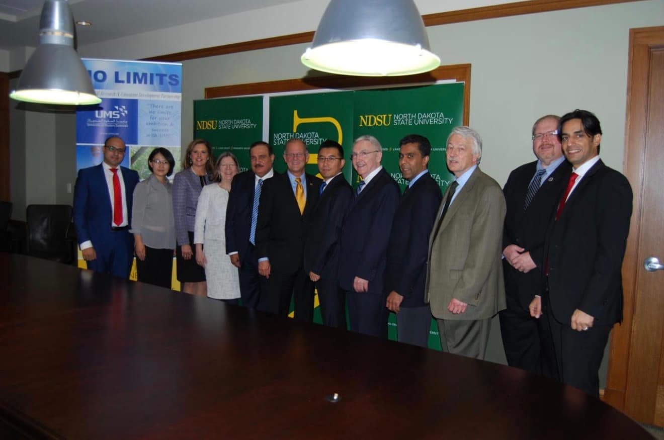 Twelve people in formal dress stand in a row in front of North Dakota State University banners and a University of Modern Sciences banner