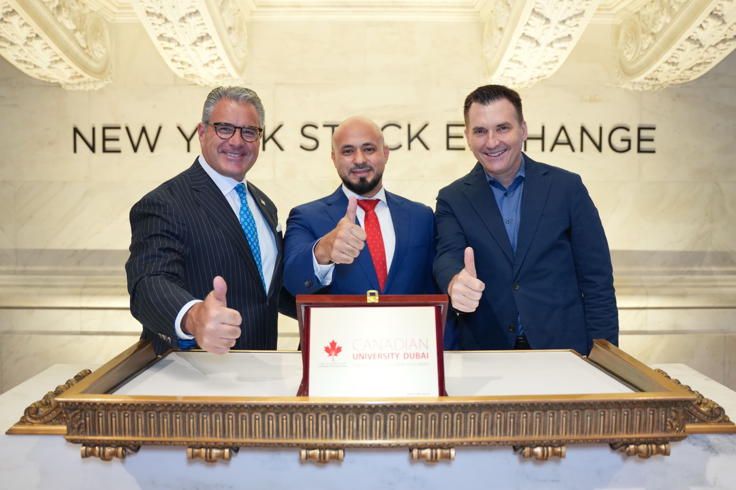 Three men at the New York Stock Exchange podium, thumbs up, behind a Canadian University Dubai plaque; Mohammad Elmoshnib stands in the middle